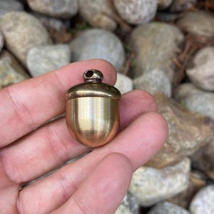 May include: A small, acorn-shaped pendant made of polished brass. The pendant has a rounded body and a cap with a small loop at the top. The brass has a warm, golden color, and the pendant is held in a hand against a background of rocks.