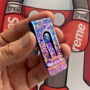 Puede incluir: Un clip para billetes multicolor con un acabado iridiscente y texturizado. El clip presenta una vibrante exhibición de azules, púrpuras y dorados, con un patrón único. El diseño incluye un recorte central para facilitar el acceso.