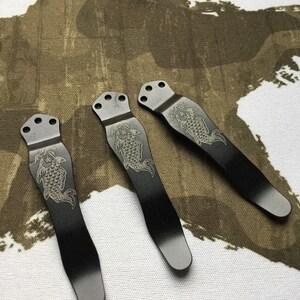May include: Three black metal pocket clips with a decorative engraved design of a fish. The clips have a rounded top and a pointed bottom.