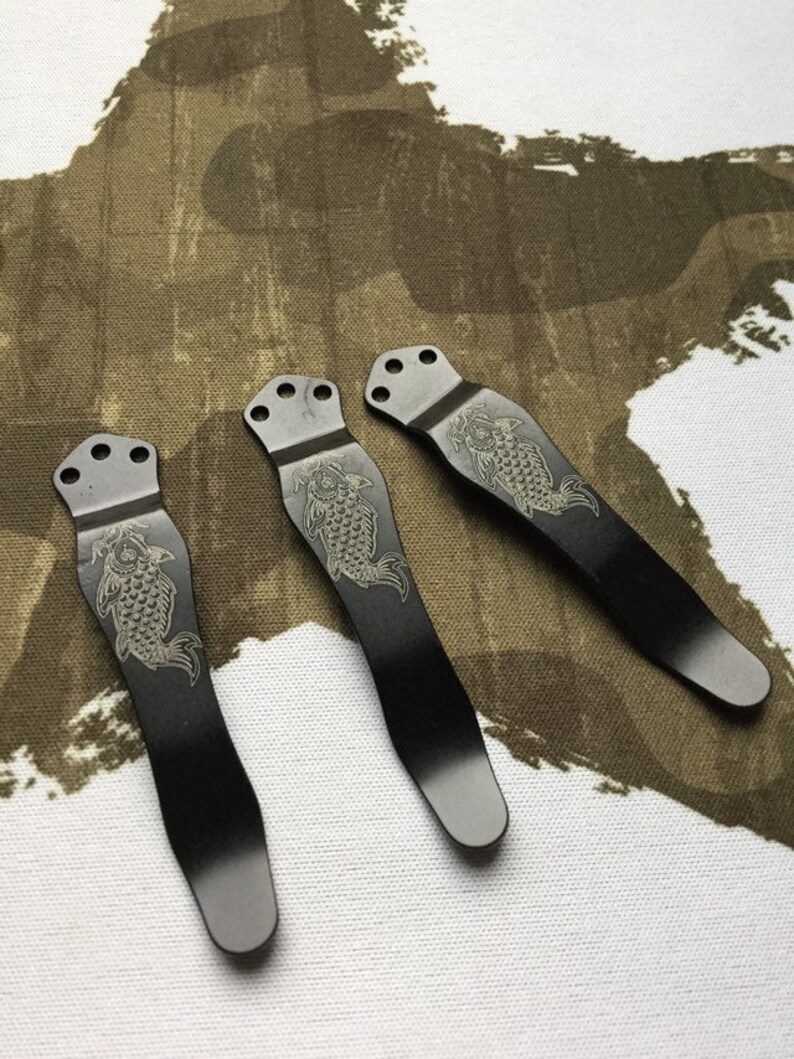 May include: Three black metal pocket clips with a decorative engraved design of a fish. The clips have a rounded top and a pointed bottom.