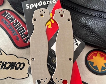 May include: Two tan knife handle scales are displayed, with a Spyderco box in the background. The scales have multiple holes and a curved design. The Spyderco box is red and black with white text. The background includes a black leather bag and patches.