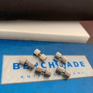 Könnte beinhalten: Fünf silberne Metallschrauben mit einem Gewindedesign, angeordnet auf einer blauen Box mit dem Text "BENCHMADE KNIFE COMPANY" in weißen Buchstaben.