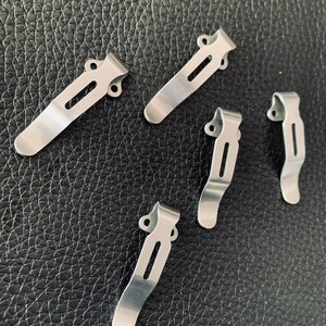K&ouml;nnte beinhalten: F&uuml;nf silberne Metall-Taschenclips mit einfachem Design. Die Clips sind so konzipiert, dass sie an einer Tasche oder einem G&uuml;rtel befestigt werden k&ouml;nnen.