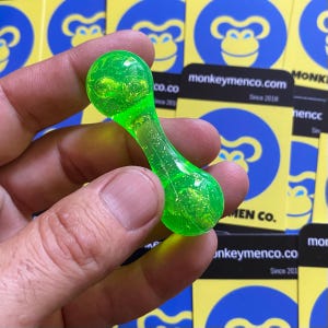 May include: A neon green, dumbbell-shaped object with a clear, textured body and rounded ends. The object is held in a hand, with a background of yellow cards featuring a monkey logo and the text "monkeymenco.com Since 2018".