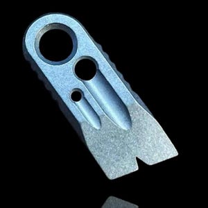 Könnte beinhalten: Ein kleines, silberfarbenes Multi-Tool mit einem Schlüsselringloch, zwei kleineren Löchern und einem spitz zulaufenden Ende. Das Werkzeug hat eine strukturierte Kante und eine Kerbe am Ende. Das Objekt ist vor einem schwarzen Hintergrund platziert.