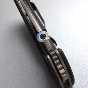 May include: Close-up of a black and silver folding knife. The knife features a silver blade, a black handle, and a pocket clip. The pivot screw has the text "LASER AUTOMAT" around the screw.