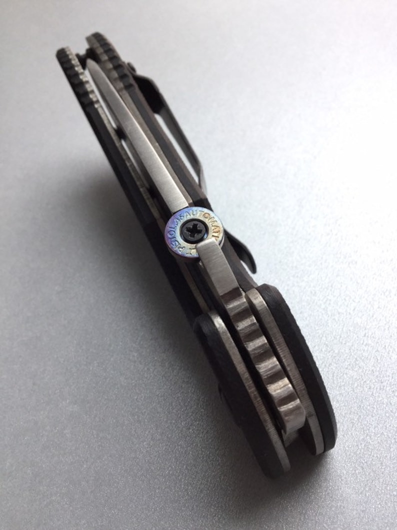 May include: Close-up of a black and silver folding knife. The knife features a silver blade, a black handle, and a pocket clip. The pivot screw has the text "LASER AUTOMAT" around the screw.