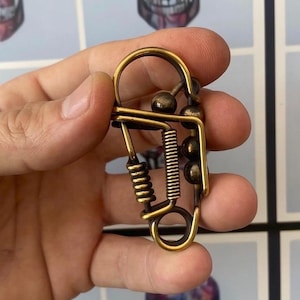 Pode incluir: Um porta-chaves de cor latão com um design abstrato único. Possui uma mola, pequenas esferas e um laço, segurado em uma mão. O porta-chaves tem uma estética vintage e provavelmente é feito de metal.