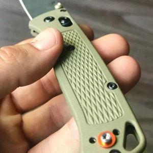 Puede incluir: Un cuchillo de bolsillo verde con una hoja negra y un mango texturizado. El cuchillo tiene un pequeño tornillo naranja en el mango.