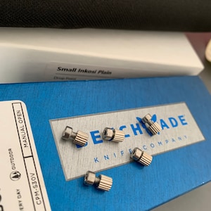 Könnte beinhalten: Nahaufnahme von fünf silbernen Schrauben auf einer blauen Box mit dem aufgedruckten Text "Benchmade Knife Company". Auf der Box steht auch der Text "Small Inkosi Plain Drop Point".