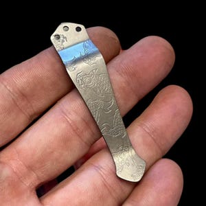 May include: A silver pocket clip with an etched design of koi fish. The clip has a slightly curved shape and a smooth, polished finish.
