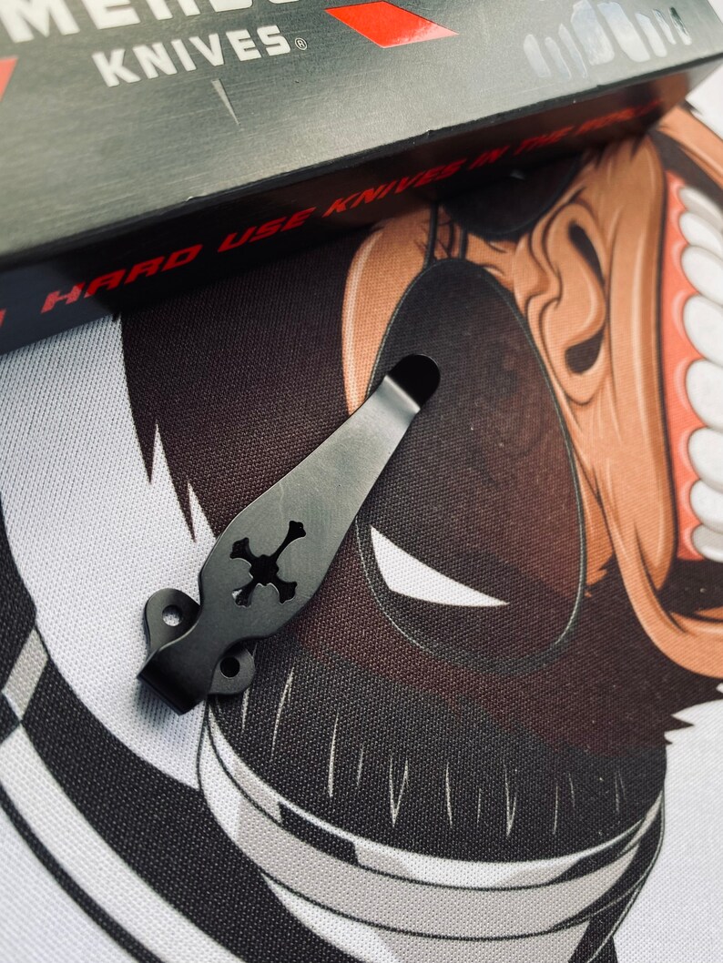 May include: Black metal pocket clip with a skull design. The clip is designed to attach to a pocket knife or other tool.