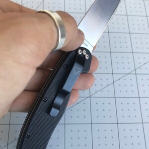 Può includere: Un coltello pieghevole nero con una lama argentata. Il coltello ha un manico strutturato e una clip da tasca nera. La lama è parzialmente aperta e la parola "Taiwan" è visibile sulla lama.
