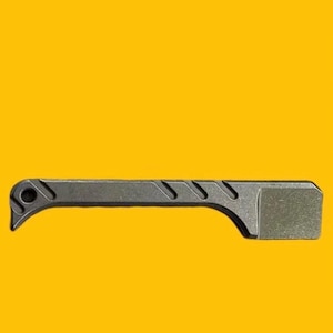 Puede incluir: Una herramienta de metal gris oscuro con un extremo rectangular y un diseño curvo y ranurado. La herramienta tiene un agujero en un extremo, sobre un fondo amarillo.