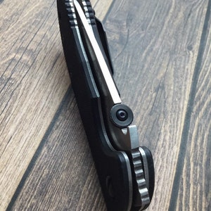 May include: A black folding knife with a silver blade and a textured handle. The knife is partially open, showing the blade and the mechanism.