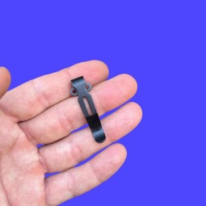 May include: A black metal pocket clip with a curved design. The clip has a rectangular slot and two small holes at the top. The clip is held in a hand against a blue background.