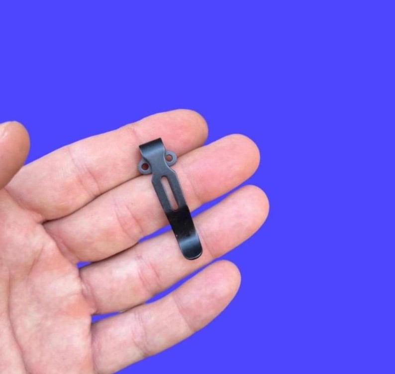 May include: A black metal pocket clip with a curved design. The clip has a rectangular slot and two small holes at the top. The clip is held in a hand against a blue background.
