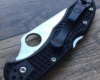 May include: A folding pocket knife with a silver-colored metal blade featuring a circular thumb hole and a black textured handle. The handle includes a black metal pocket clip with a small spider logo and several silver-colored screws.