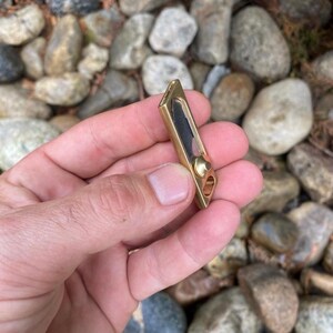 May include: A small, rectangular brass pocket knife with a black blade. The knife has a rounded end with a small hole and a brass rivet. The knife is held in a hand against a background of rocks.
