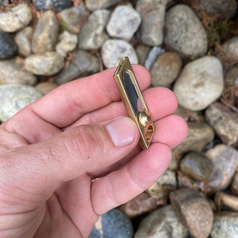 May include: A small, rectangular brass pocket knife with a black blade. The knife has a rounded end with a small hole and a brass rivet. The knife is held in a hand against a background of rocks.