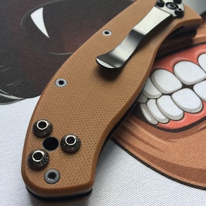 May include: A tan pocket knife with a black clip and a textured handle. The knife has a black blade and a black pocket clip.