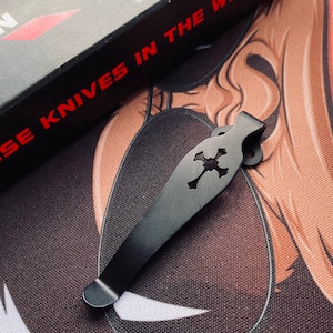 May include: Black metal pocket clip with a skull and crossbones design. The clip is designed to attach to a pocket knife.