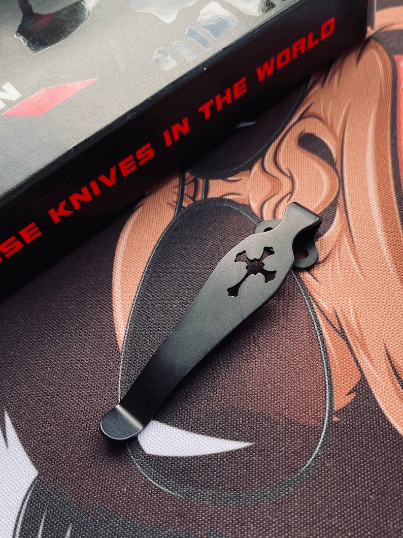 May include: Black metal pocket clip with a skull and crossbones design. The clip is designed to attach to a pocket knife.