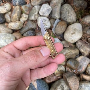 May include: A small, brass pocket knife with a silver blade. The knife is open, revealing the sharp edge. The brass handle has a rectangular shape with rounded edges and a small hole at the end. The knife is held against a background of rocks.