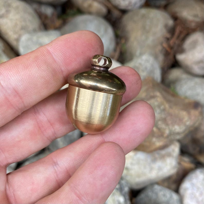 May include: A small, acorn-shaped container made of polished brass. The container has a rounded cap with a small loop at the top. The acorn is held in a hand, with a blurred background of rocks and foliage.