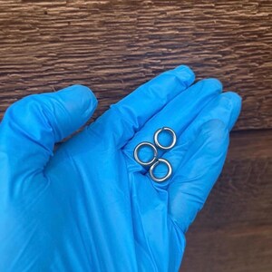 Puede incluir: Tres pendientes de aro plateados. Los pendientes circulares pequeños son de metal. Se muestran en una mano enguantada de azul sobre un fondo de madera marrón.