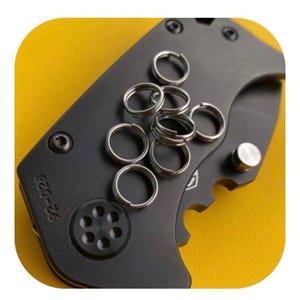 Op de afbeelding: Een zwart zakmes met zilveren sleutelringen op een gele ondergrond. Het mes heeft een gedeeltelijk gekarteld lemmet en een zilveren duimknop. De sleutelringen zijn gemaakt van zilverkleurig metaal.