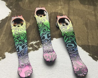 May include: Three metal pocket clips for knives, each featuring a vibrant, multi-coloured gradient pattern with swirling, topographic-like lines in shades of green, blue, purple, and pink. Each clip has a circular mounting hole at the top end.