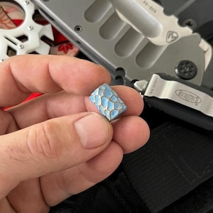 K&ouml;nnte beinhalten: Eine kleine, rechteckige Perle mit einem hellblauen und goldenen Wabenmuster. Die Perle wird in einer Hand gehalten, mit einem silbernen und schwarzen Messer und anderen Werkzeugen im Hintergrund.