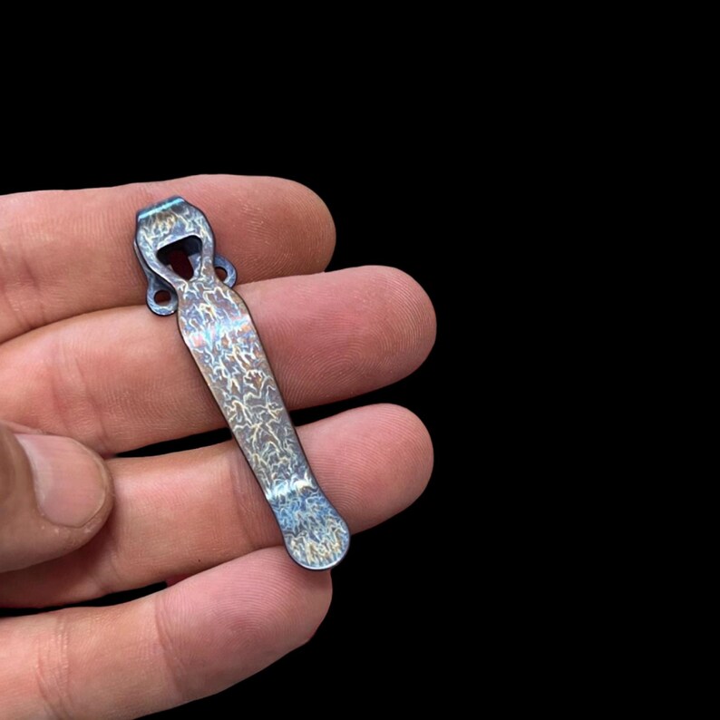 May include: A blue and silver pocket clip with a unique patterned design. The clip is made of metal and has a rounded end.