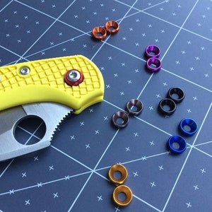 Op de afbeelding: Assortiment van kleurrijke metalen ringen in verschillende kleuren, waaronder goud, blauw, zwart, paars en koper. De ringen zijn gerangschikt in rijen op een zwarte rasterachtergrond.