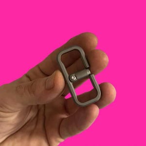 Könnte beinhalten: Ein kleiner, rechteckiger, silberfarbener Drehverschluss mit einem zentralen Drehmechanismus. Der Verschluss wird vor einem leuchtend pinkfarbenen Hintergrund gehalten. Der Verschluss ist zum Drehen und Verbinden von Gegenständen konzipiert.