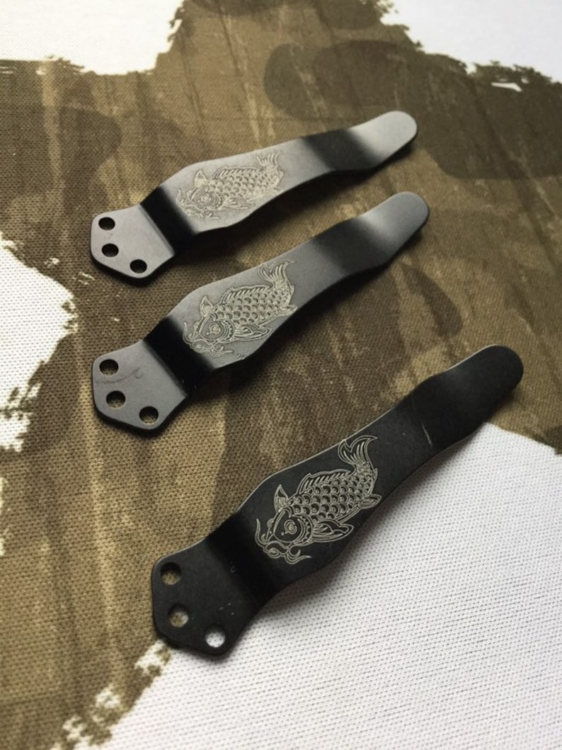 May include: Three black metal knife scales with a decorative koi fish design. The scales have three holes for attaching to a knife.