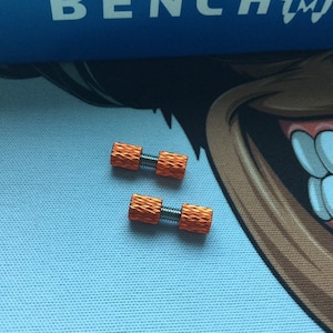 Könnte beinhalten: Zwei orangefarbene Metallschrauben mit strukturiertem Design. Die Schrauben haben eine silberne Mitte und liegen auf einer hellblauen Oberfläche. Das Wort "BENCHMADE" ist im Hintergrund sichtbar.