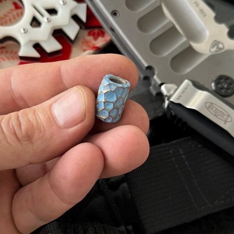 K&ouml;nnte beinhalten: Eine kleine, blau-silberne Perle mit einem Wabenmuster. Die Perle ist zylindrisch und scheint aus Metall zu sein. Die Perle wird in der Hand einer Person gehalten, mit einem teilweise sichtbaren Messer und anderen Objekten im Hintergrund.