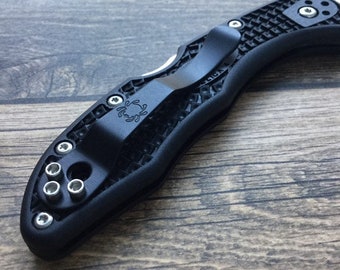 May include: A black folding knife handle featuring a textured grip pattern, a matte black pocket clip with a small spider logo, and several silver-toned metal screws securing the clip and handle components together in a compact design.