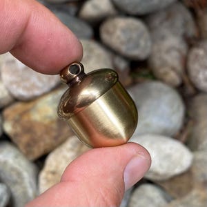 May include: A small, acorn-shaped pendant made of polished brass. The acorn has a rounded body and a detailed cap with a small loop for a chain. The brass has a warm, golden hue, and the pendant is held between two fingers.