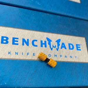 Könnte beinhalten: Ein goldfarbener Metallstift mit dem Logo von Benchmade Knife Company mit einem Schmetterlingsdesign.