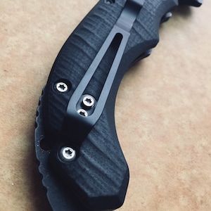 Könnte beinhalten: Ein schwarzes Taschenmesser mit einem strukturierten Griff und einem Deep-Carry-Taschenclip. Der Clip ist mit drei Schrauben befestigt.
