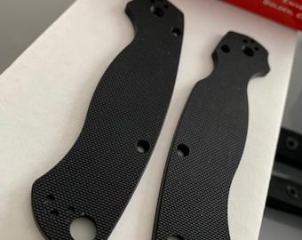 May include: Two black plastic knife scales with holes for mounting hardware. The scales are textured and have a Spyderco logo on the box in the background.