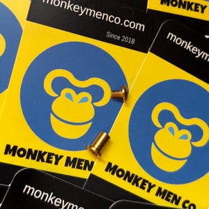 Puede incluir: Tarjetas de visita con un logotipo de mono amarillo y azul y el texto "MONKEY MEN CO." y "monkeymenco.com Since 2018". Un pequeño tornillo dorado y un tubo de metal dorado están encima de las tarjetas.