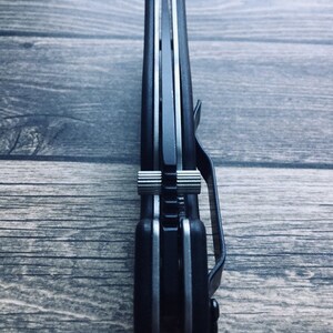 May include: A black folding knife with a silver blade and a pocket clip. The knife is closed, showing the side profile with a textured handle and a silver accent. The knife is resting on a wooden surface.