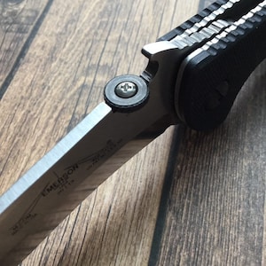 K&ouml;nnte beinhalten: Ein schwarzes und silbernes Klappmesser mit einem strukturierten Griff und einer teilweise ge&ouml;ffneten Klinge. Die Klinge tr&auml;gt die Gravuren "EMERSON" und "MADE IN USA".
