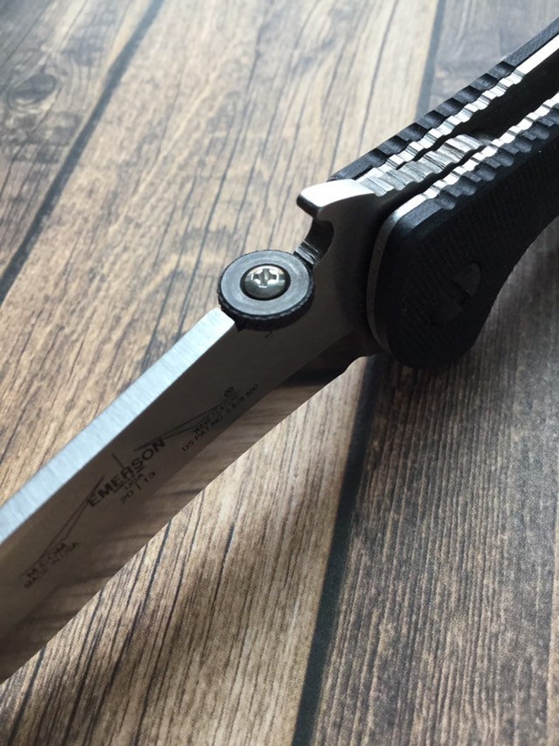 K&ouml;nnte beinhalten: Ein schwarzes und silbernes Klappmesser mit einem strukturierten Griff und einer teilweise ge&ouml;ffneten Klinge. Die Klinge tr&auml;gt die Gravuren "EMERSON" und "MADE IN USA".