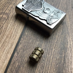 Puede incluir: Un encendedor plateado con un logotipo de Harley Davidson y un diseño de águila. El encendedor está hecho en los Estados Unidos. También hay un pequeño spinner de metal dorado en la imagen.