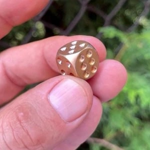 Puede incluir: Un pequeño dado dorado con bordes redondeados. El dado tiene hendiduras circulares que representan números, con un total de seis caras. El dado está sostenido en la mano de una persona, con un fondo verde borroso.